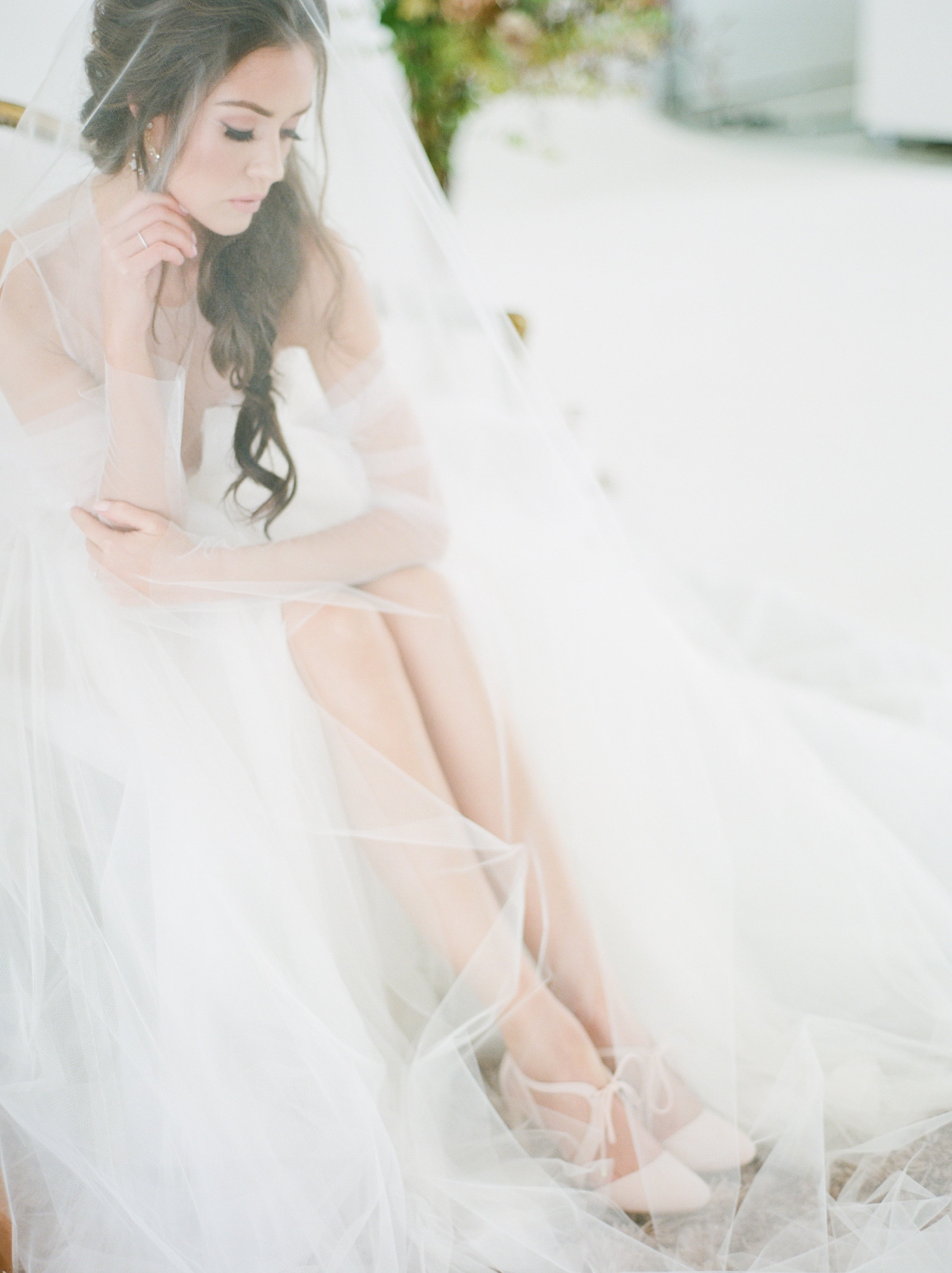 Serene bride sitting gracefully under her voluminous tulle veil, adjusting her elegant bridal shoes. The soft lighting and delicate details of her dress create a dreamy and ethereal wedding day look.