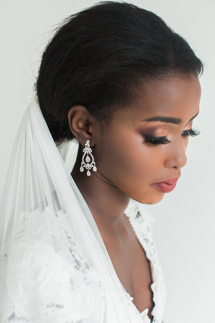 Close-up of a radiant bride adorned with dazzling chandelier earrings and a classic white veil. The intricate lace detailing of her gown and her elegant makeup highlight her sophisticated wedding day look.